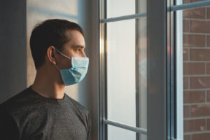 A man looks out the window while wearing a mask. This could represent the isolation of quarantine that online depression therapy in New Jersey can offer support with. Contact a therapist in South Jersey to learn more about therapy for anxiety in South Jersey and online therapy in New Jersey. 08085