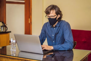 A man wearing a mask types on a laptop. This could represent a client in a session of online depression therapy in New Jersey. Learn more about online therapy in New Jersey by contacting a therapist in South Jersey to learn more about therapy in Swedesboro, NJ. 08085