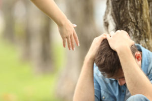A teen holds their head as they sit against a tree while a helping hand reaches towards them. This could represent the support a teenage therapist in Swedesboro, Nj can offer with online therapy for teens in New Jersey. Learn more by searching "teenage therapist near me in washington, nj" today! 08085