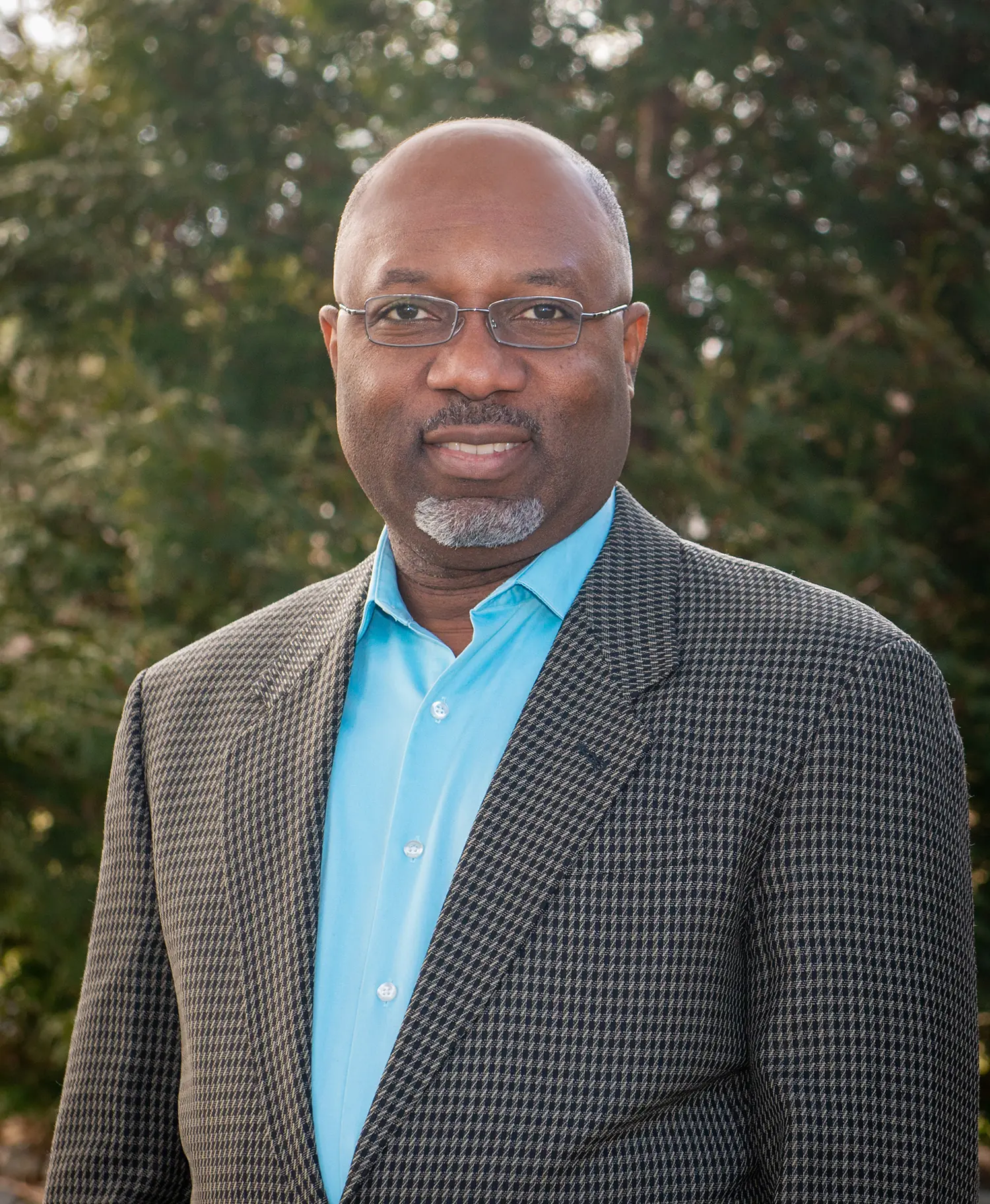 a man named Yvans Jazon in a suit and glasses standing in front of trees